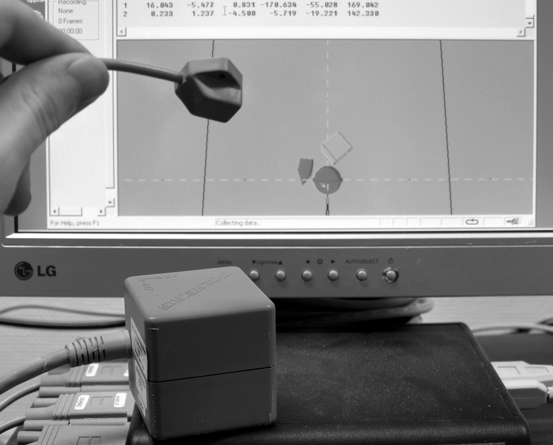 Figur 7: Et elektromagnetisk sensorsystem i bruk. Kuben nederst i bildet er senderen som setter opp det elektromagnetiske feltet. En av sensorene holdes i hånden, og en grafisk fremstilling av sensorens posisjon og rotasjon kan ses på skjermen i bakgrunnen.